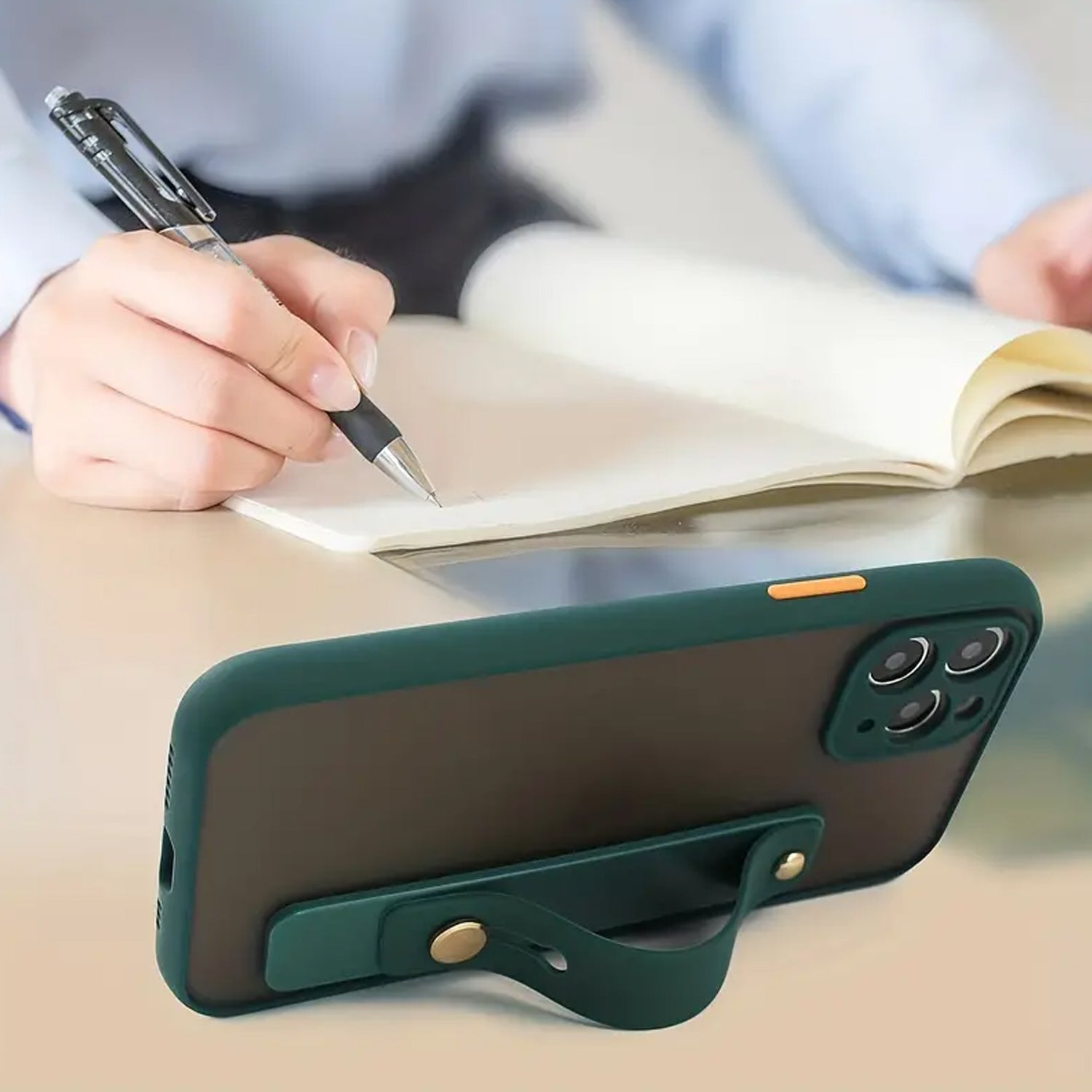 A dark green phone with a matching silicone grip acting as a kickstand, with a person writing in a notebook in the