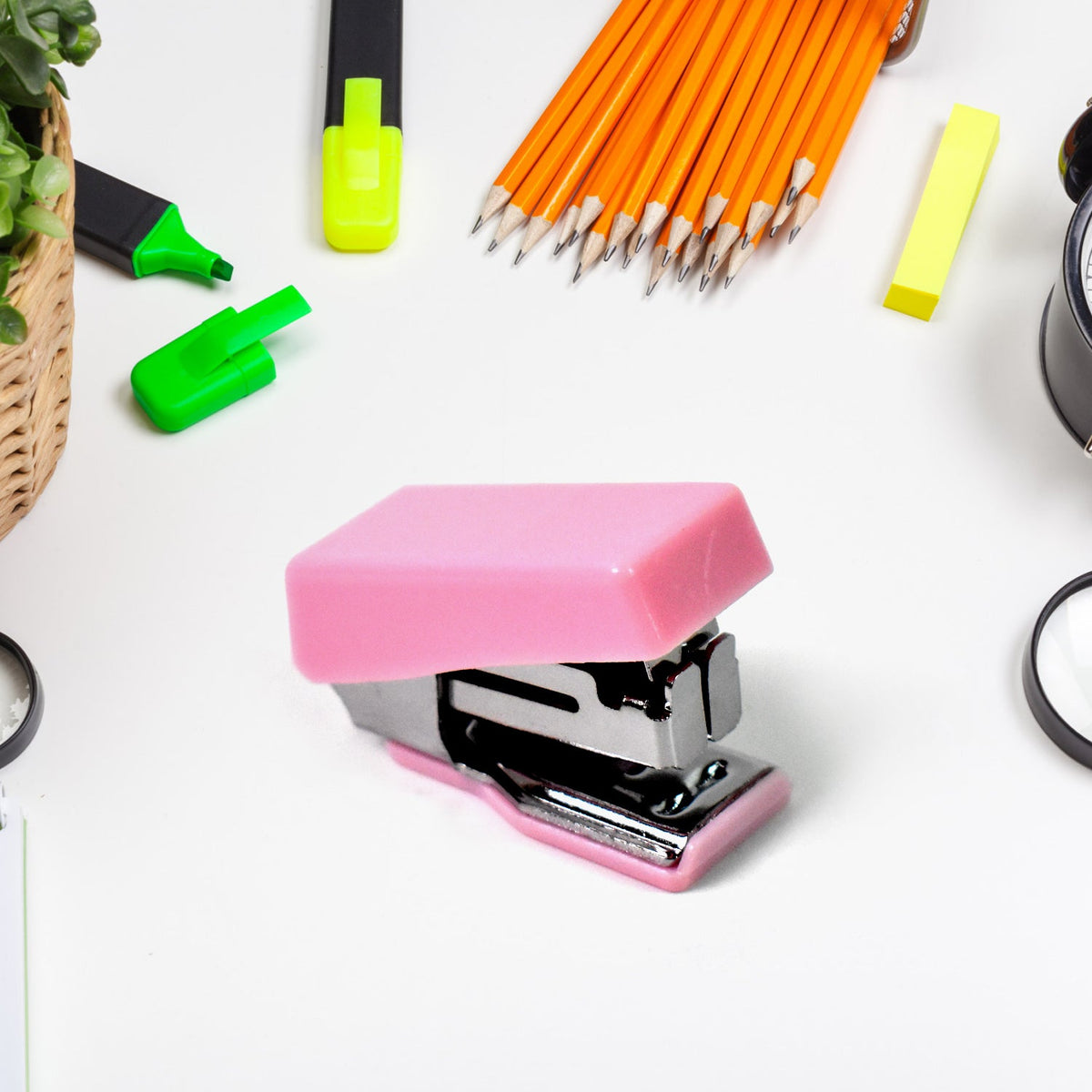A close-up of a pink mini stapler on a white desk surrounded by yellow pencils and green highlighters.