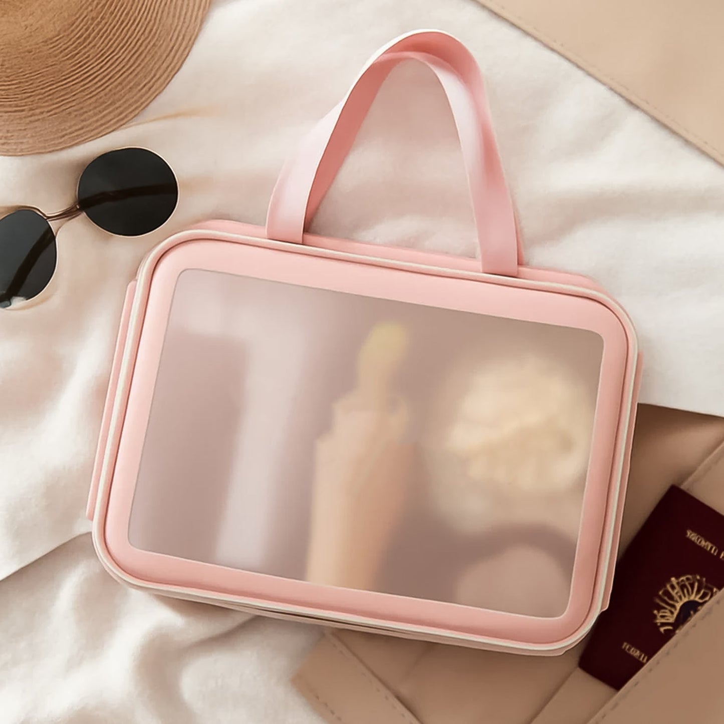 A large pink waterproof travel bag with a clear panel lies on a bed next to a straw hat and sunglasses.