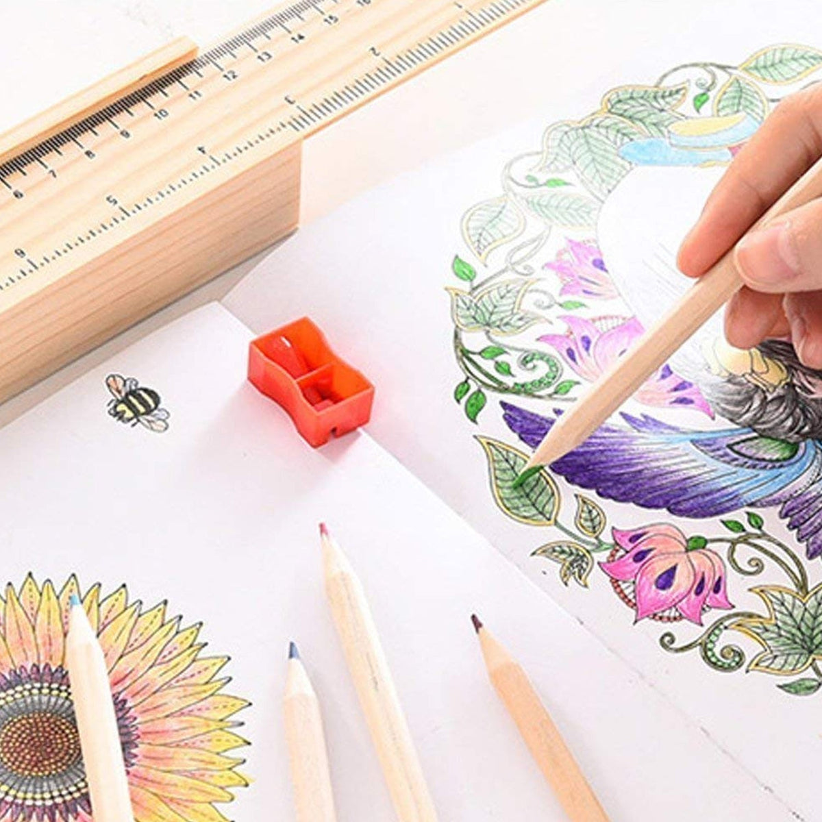 A hand colors a detailed floral and bird design in a coloring book with a light wood pencil, next to a ruler and sharpener.