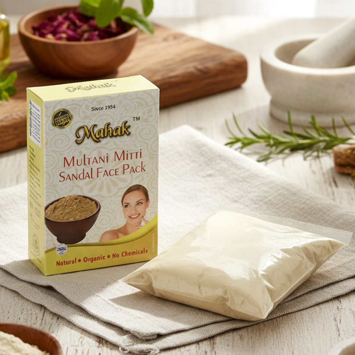 Mahak Multani Mitti Sandal Face Pack box and a clear bag of powder on a light grey napkin, surrounded by herbs and bowls.