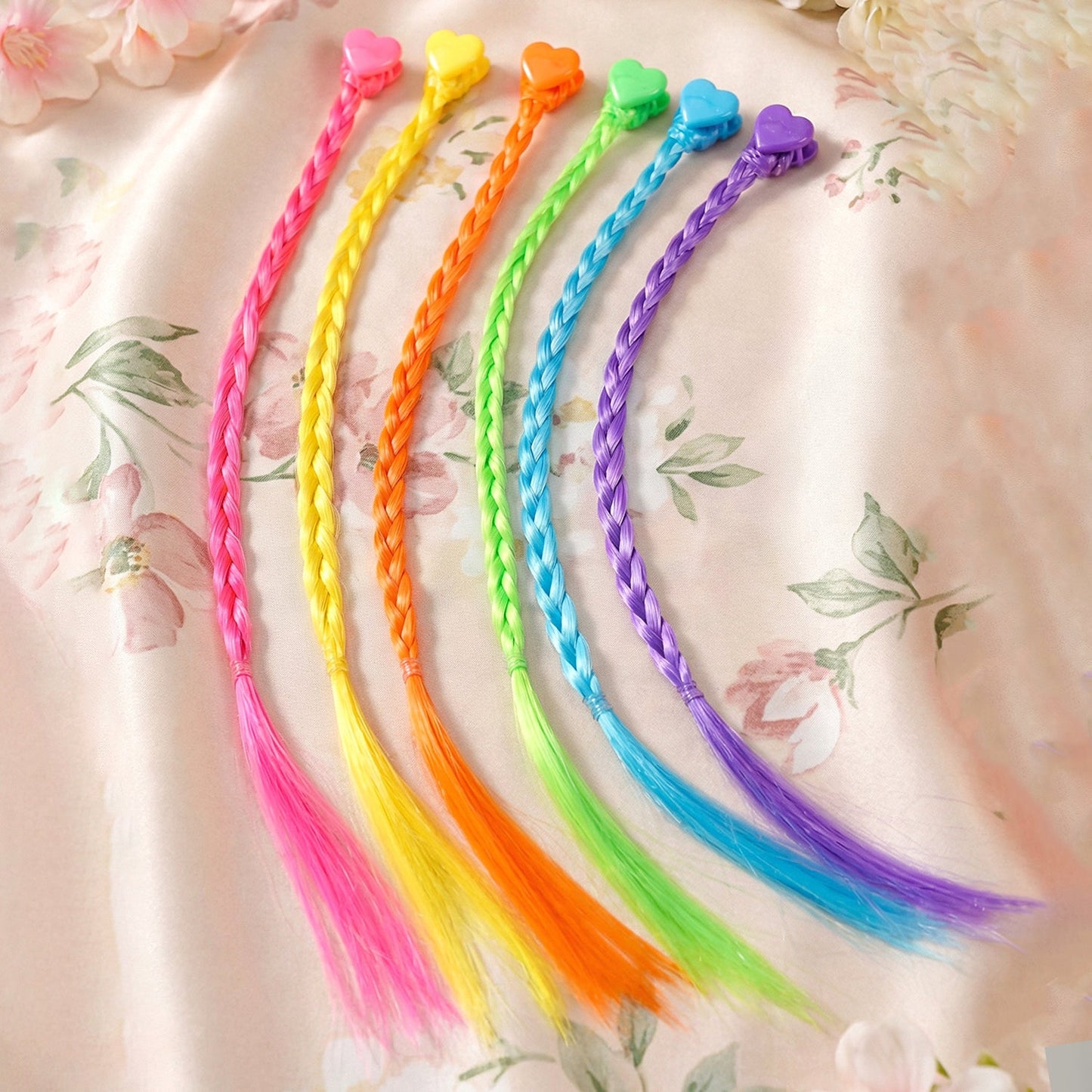 Six brightly colored braided hair extensions with heart-shaped clips are displayed on a floral patterned fabric.