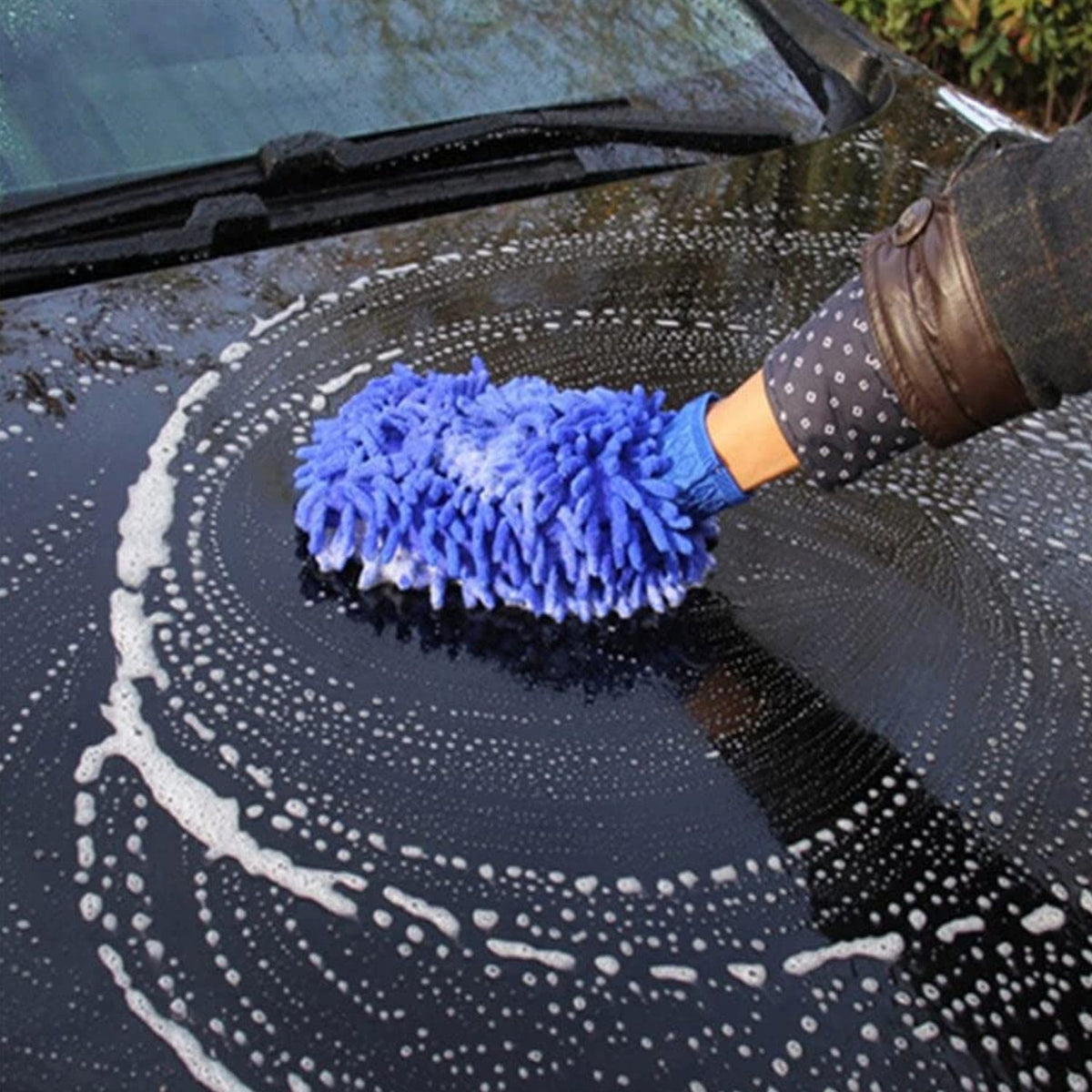 A hand wearing a blue microfiber wash mitt creates circular soap patterns on a black car hood.