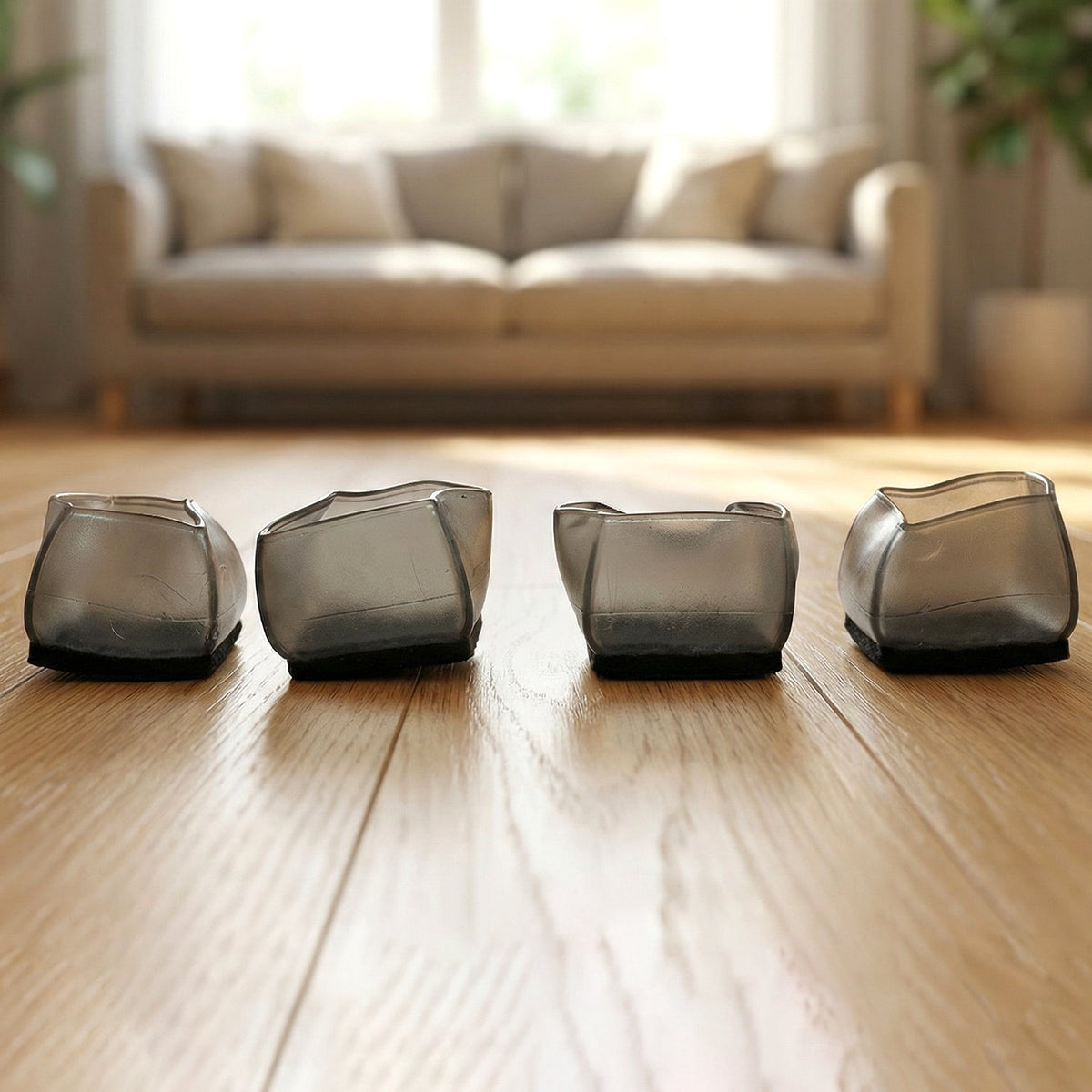Four dark grey silicone furniture leg caps are lined up on a wooden floor, with a blurred living room in the background.