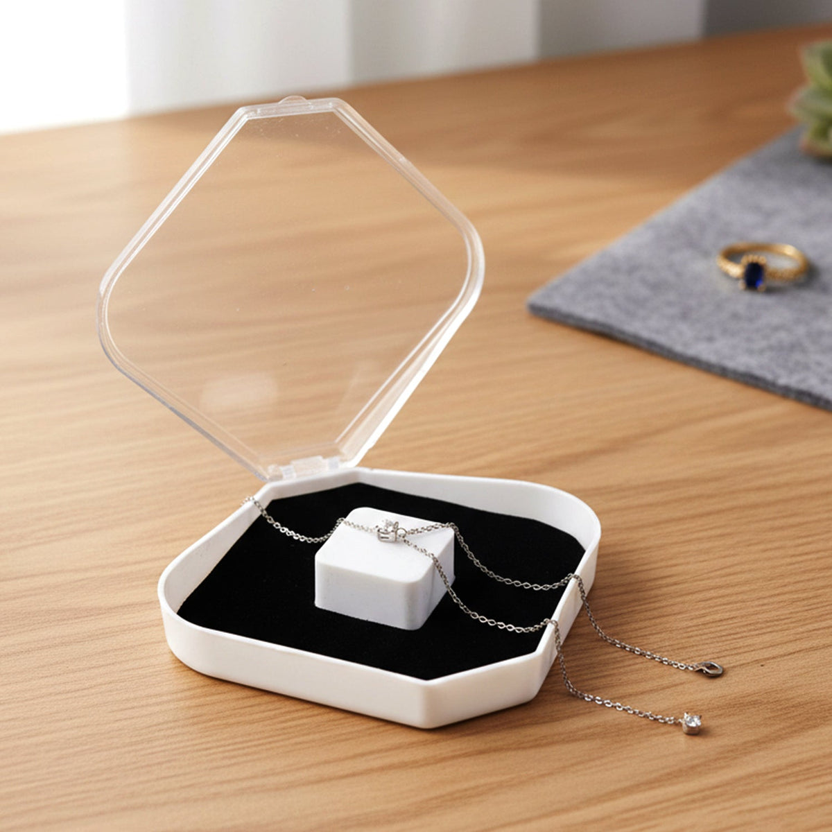 An open clear and white jewellery box with a silver necklace on a black cushion, next to a ring on a grey mat.