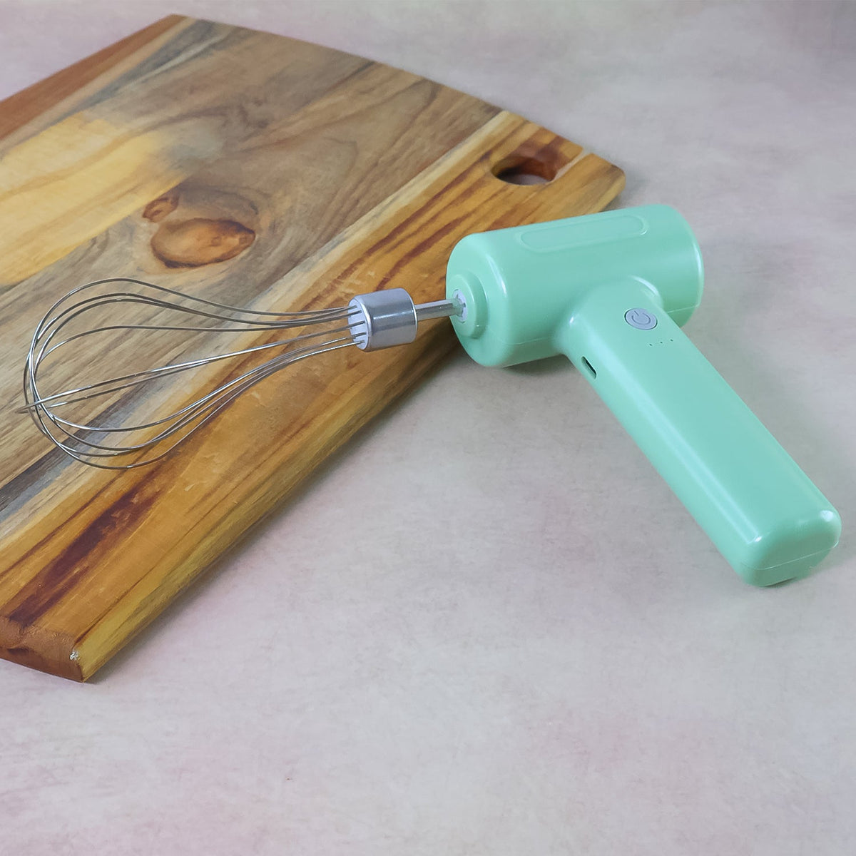 Light green cordless electric whisk with its attachment resting on a brown counter next to a wooden cutting board.