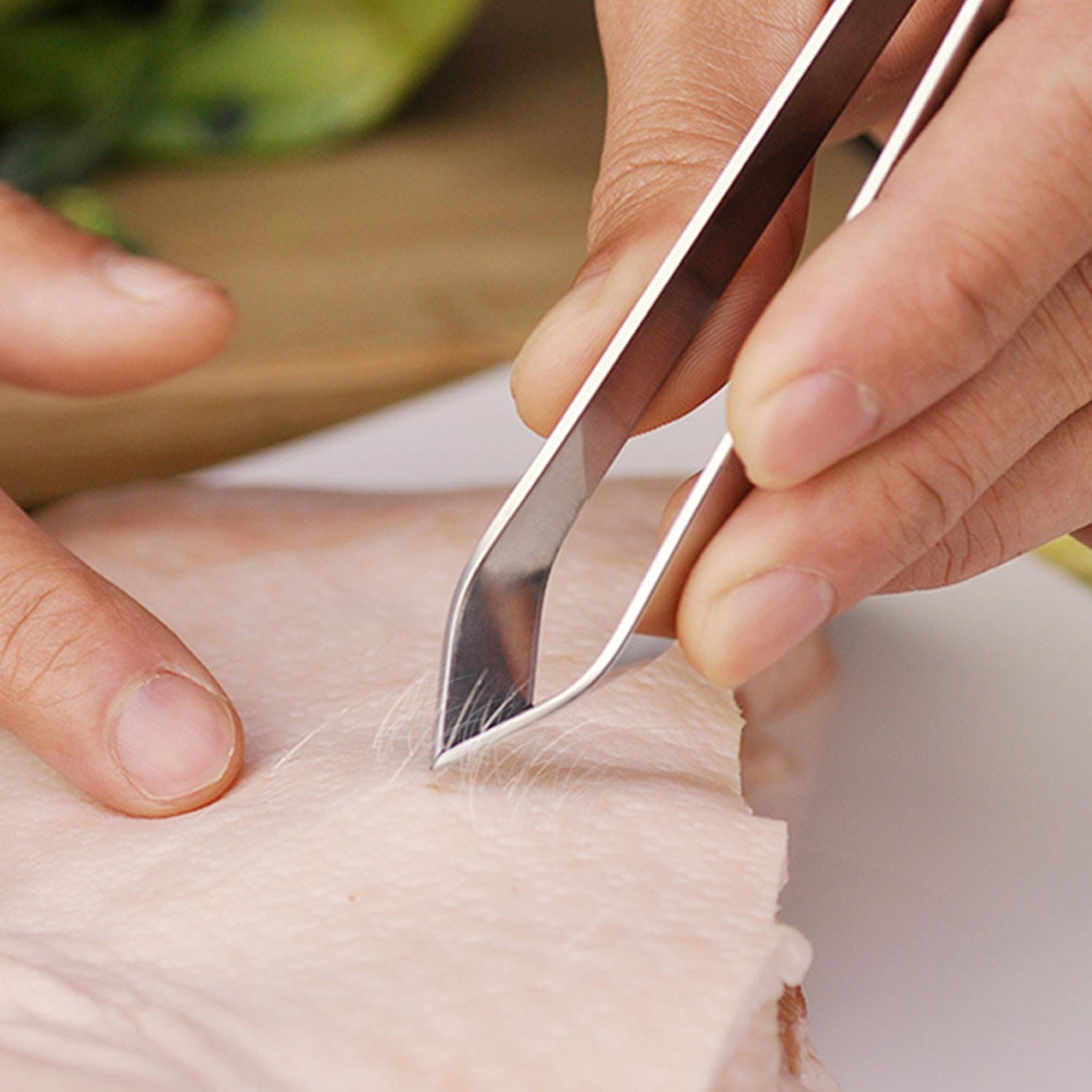 A close-up of hands using silver stainless steel tweezers to remove hair from a piece of raw meat.