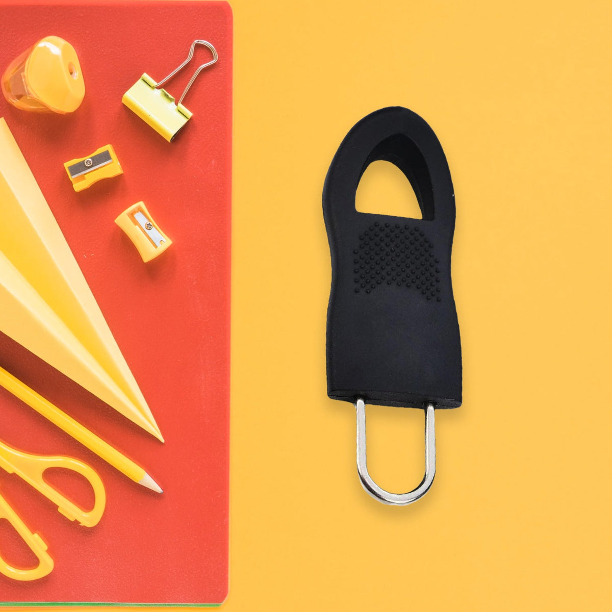 A black rubber zipper pull tab with a silver metal loop is on a yellow surface next to a red notebook.