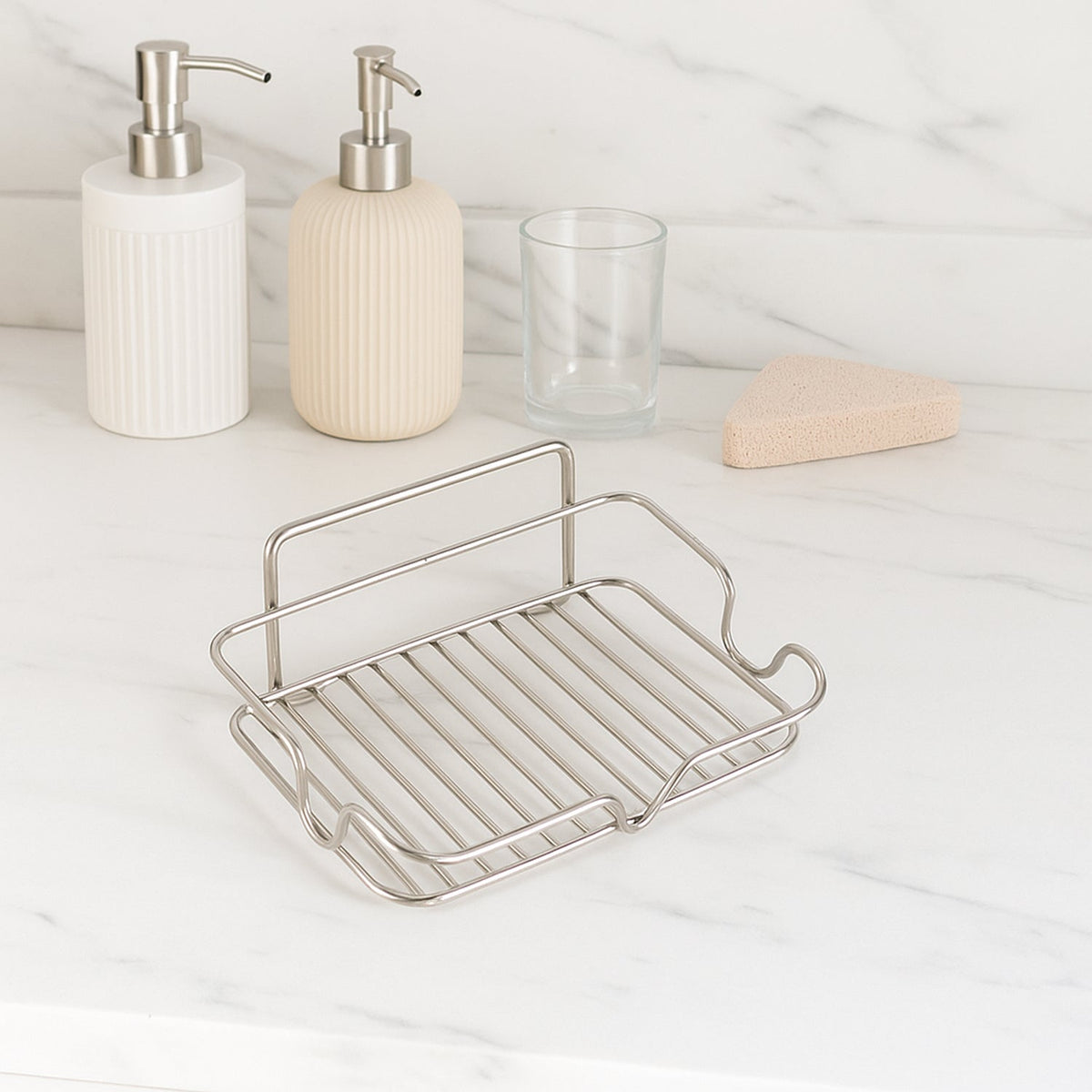 A stainless steel wire soap dish sits on a white marble counter with two soap dispensers and a glass in the background.