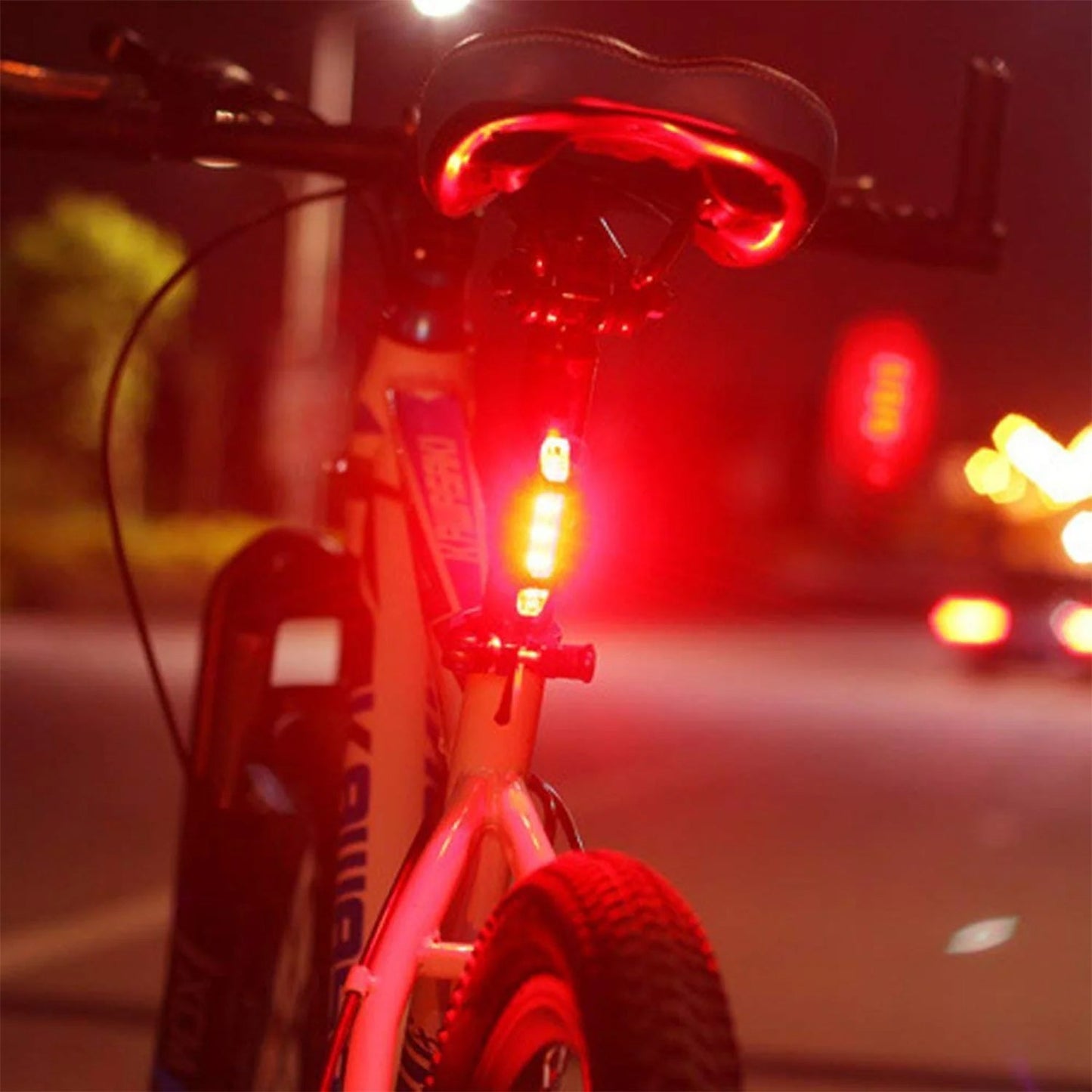 A red LED bicycle tail light illuminates the back of a bicycle at night, with blurred city lights in the background.