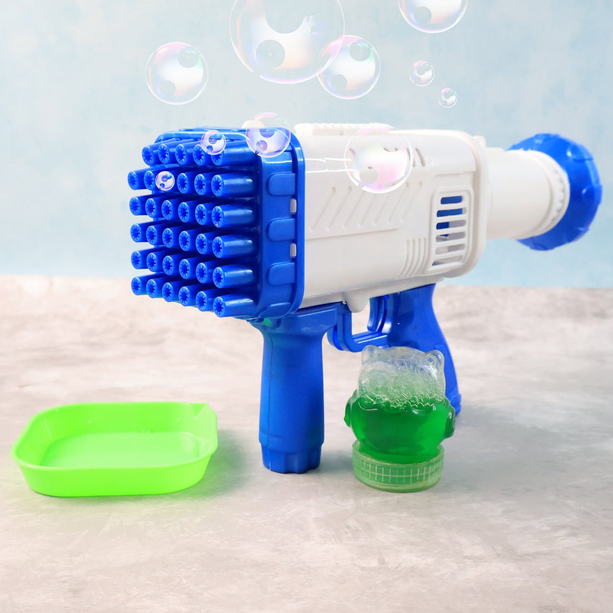 A white and blue 32-hole bubble gun, a green bubble solution tray, and a bottle of solution on a grey surface.