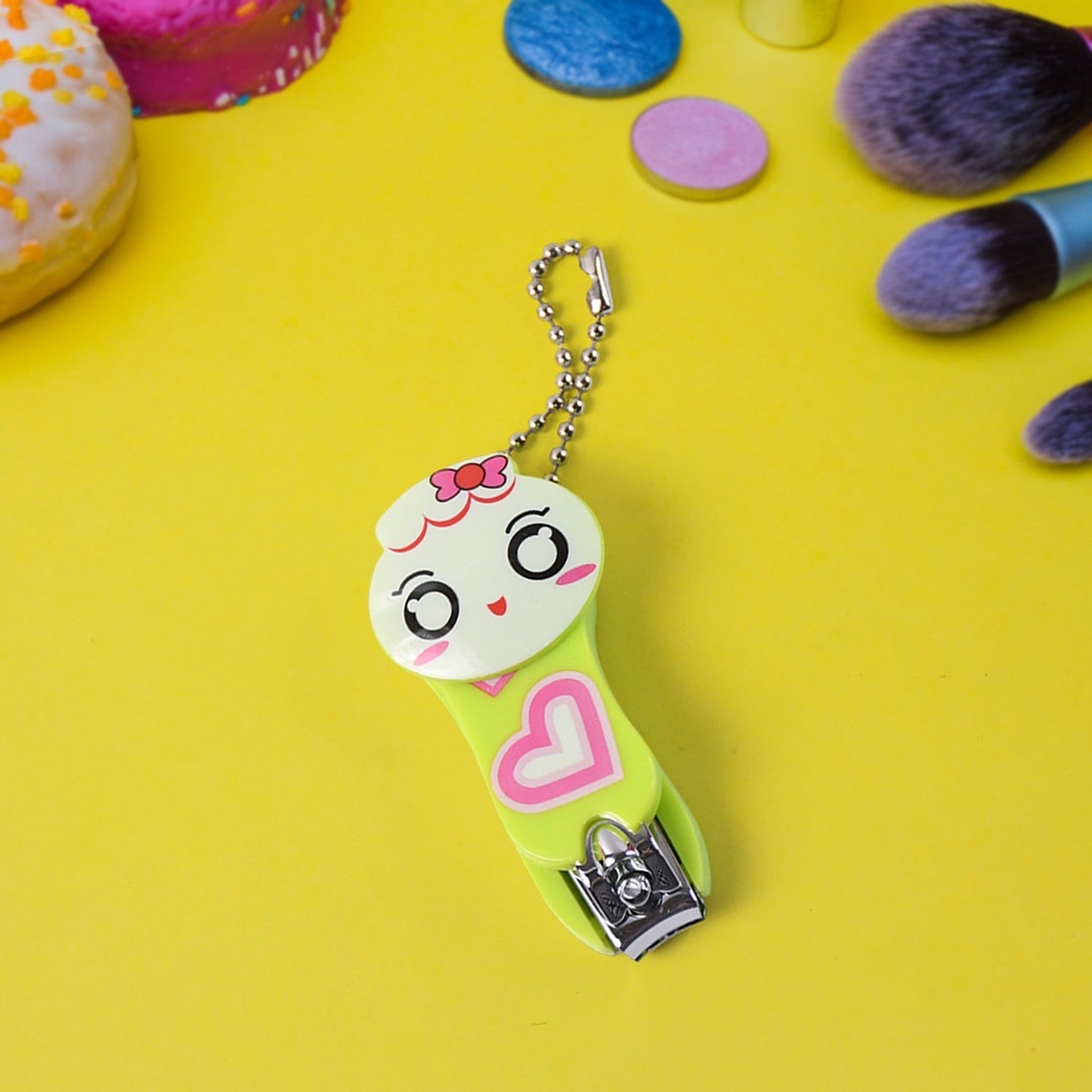 A green and white cartoon character nail clipper with a pink heart and a silver ball chain, on a bright yellow surface with