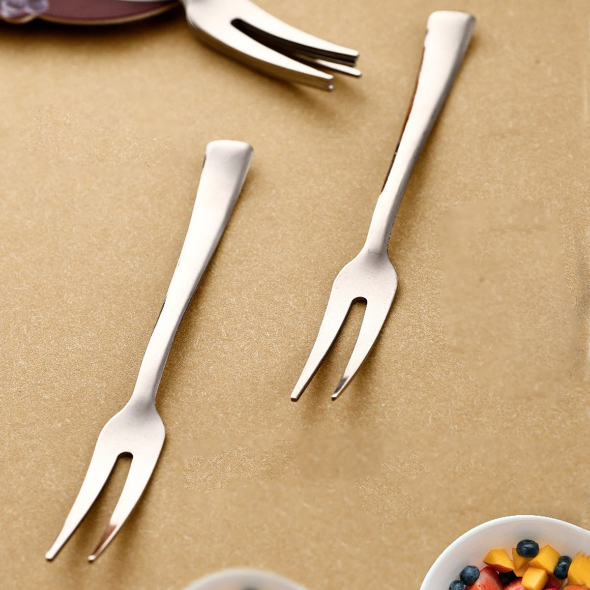Two silver stainless steel fruit forks with two tines each, resting on a light brown surface with fruit in a bowl.