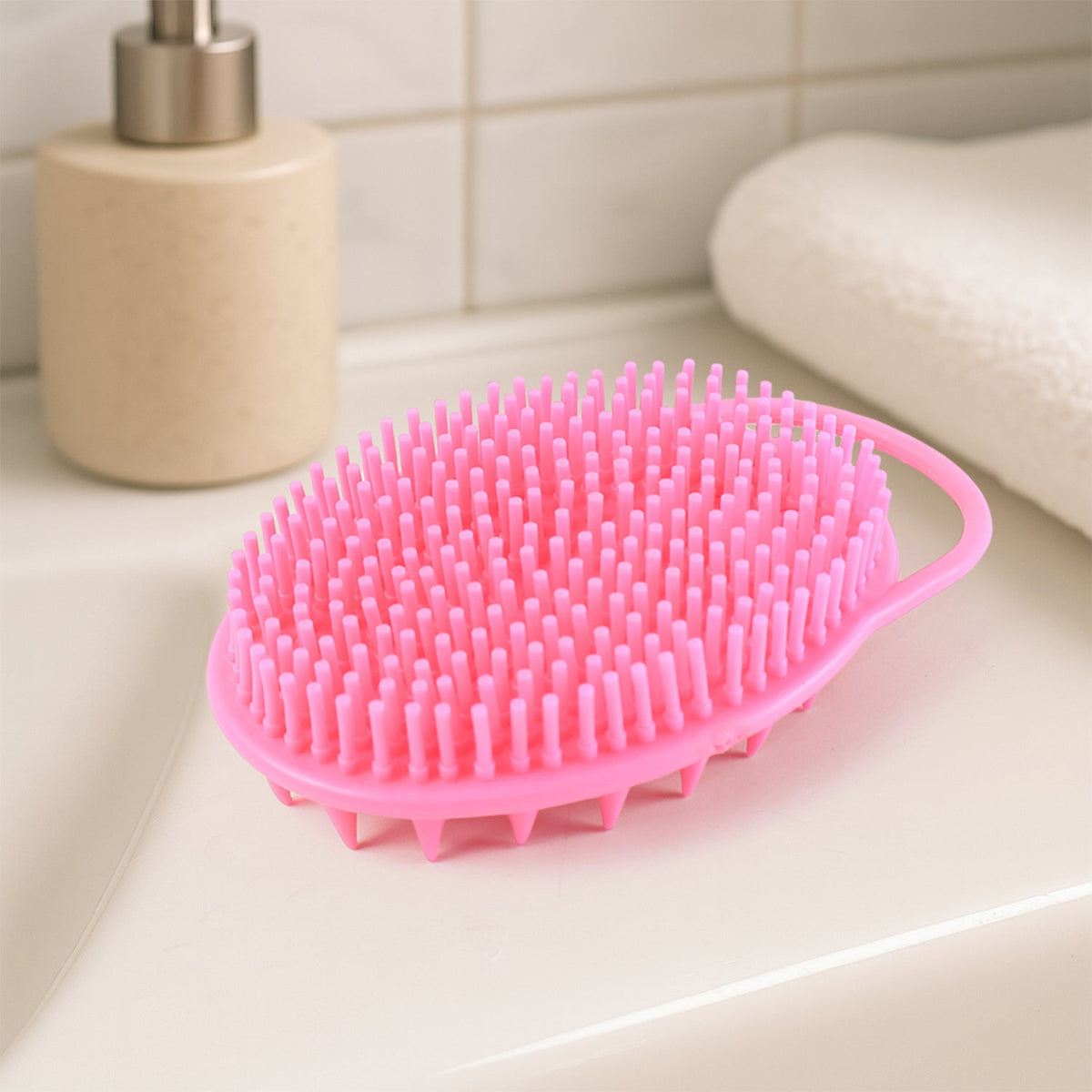 A pink silicone body brush with spiky and soft bristles sits on a white bathroom counter next to a soap dispenser and a