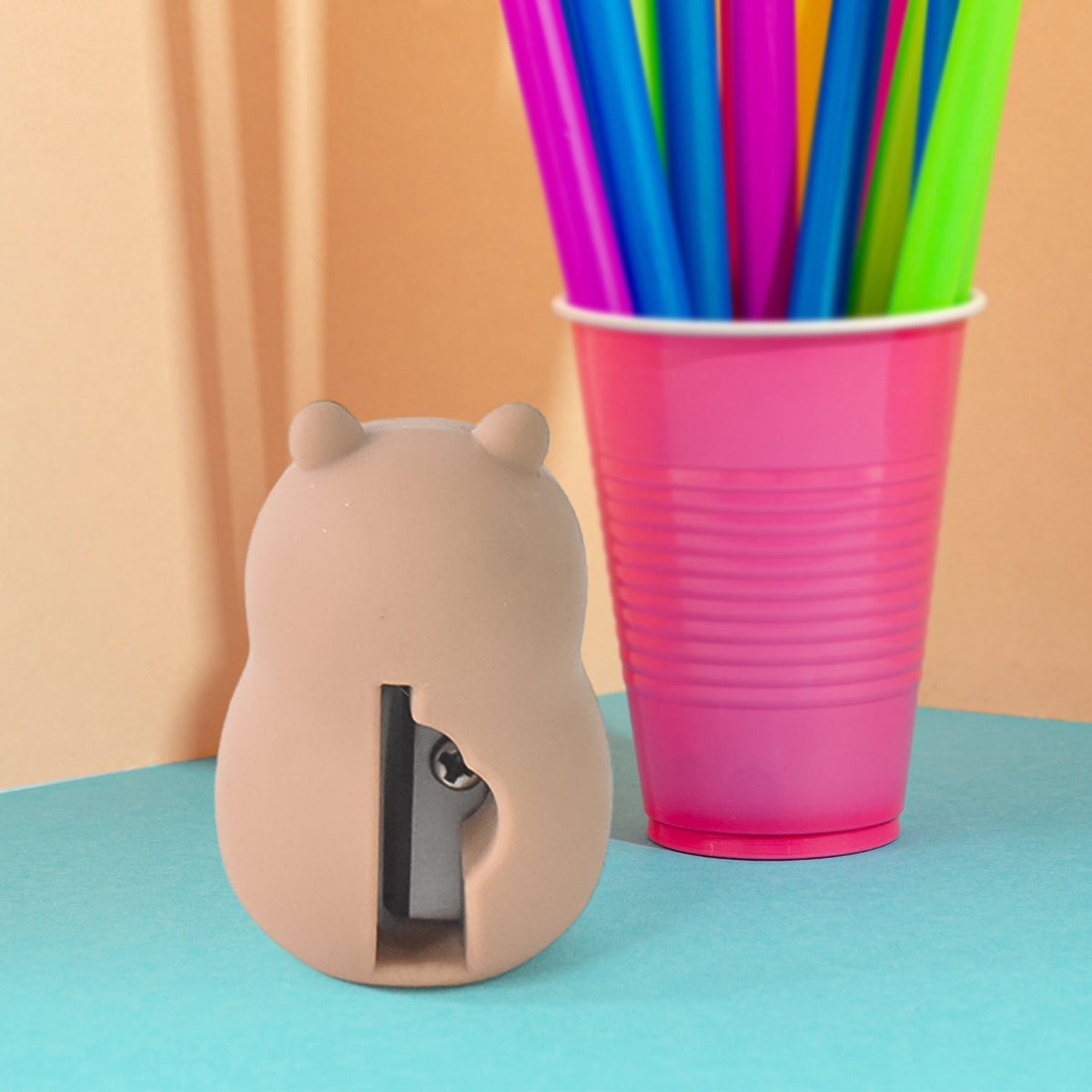 A capybara-shaped pencil sharpener, brown with a pink drink, sits next to a pink cup of colorful straws.