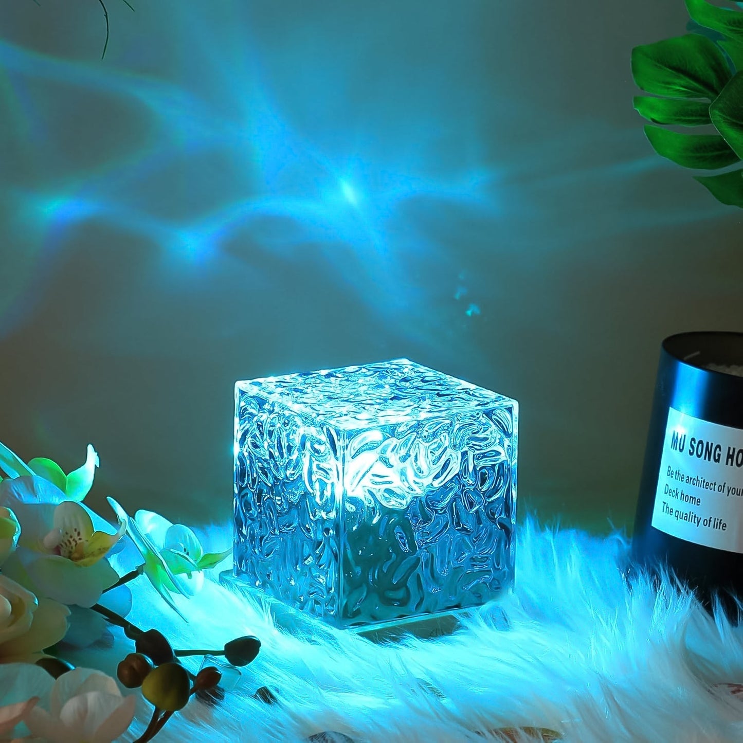 A crystal cube lamp projecting blue ripples, sitting on a white fluffy surface next to a plant and a black candle.