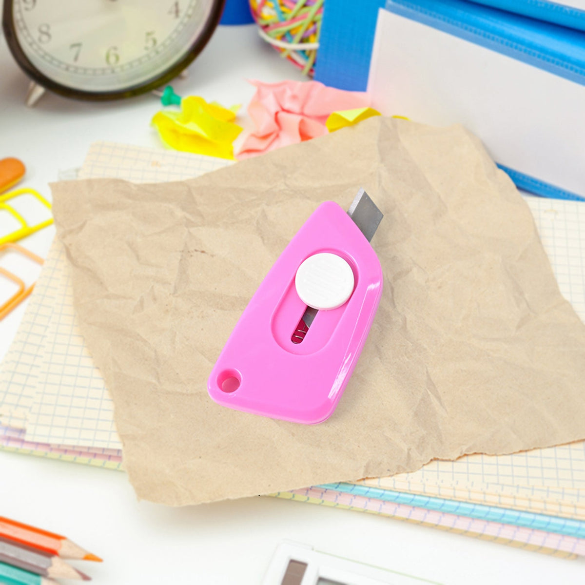 A pink retractable utility knife on crumpled brown paper, with an alarm clock and office supplies in the background.