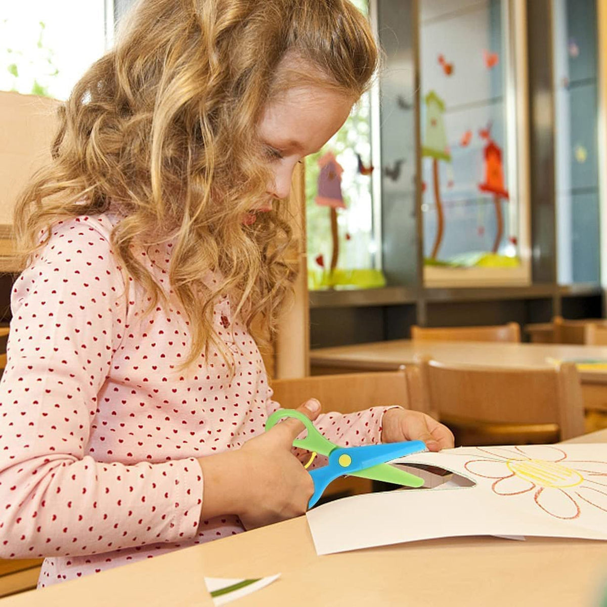 A young girl with long curly hair cuts a flower shape from white paper with blue and green plastic scissors.