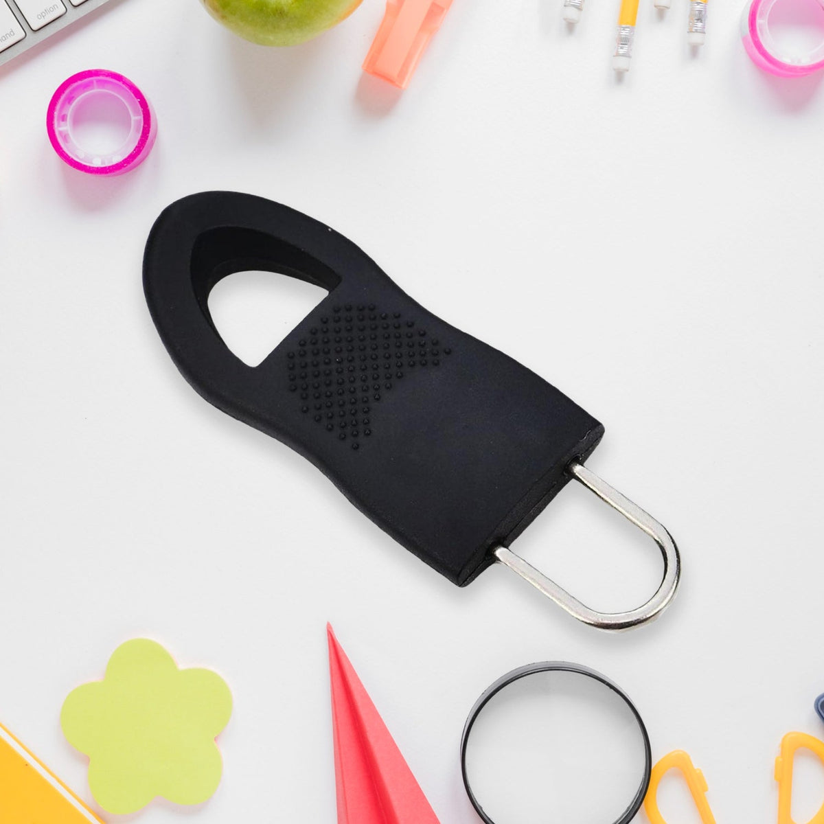 A black rubber zipper pull tab with a silver metal loop is on a white surface surrounded by various office supplies.
