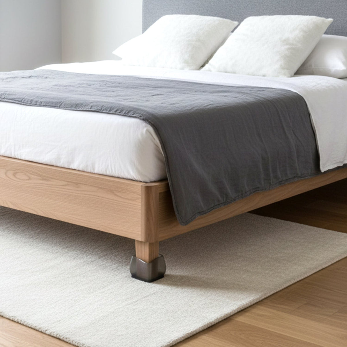 A dark grey silicone cap is on a wooden bed leg, with a white rug and bed with grey throw in the background.
