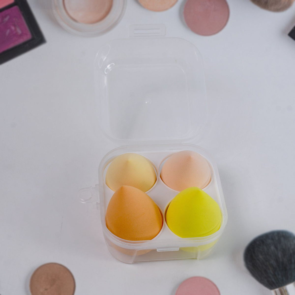 A clear plastic case holds four colorful makeup sponges, surrounded by various makeup products on a white surface.