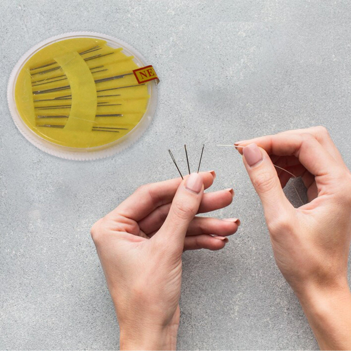 Hands holding three sewing needles and a thread, with a yellow needle disc in the background.