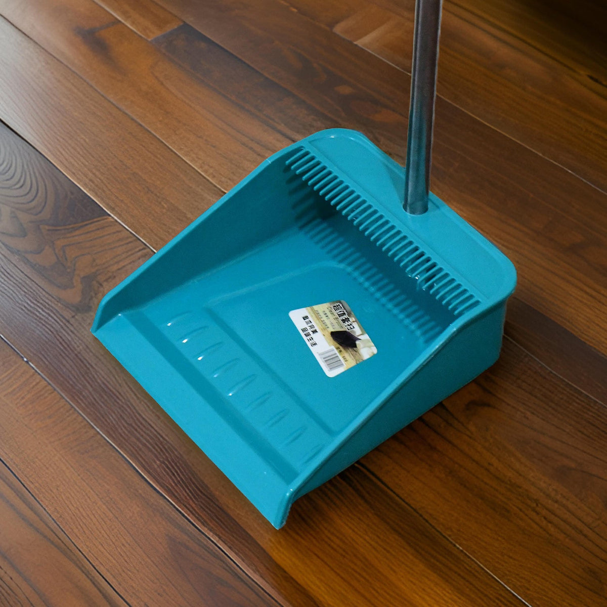 A close-up of a teal dustpan with a long silver handle on a dark wood floor, showing its textured surface.