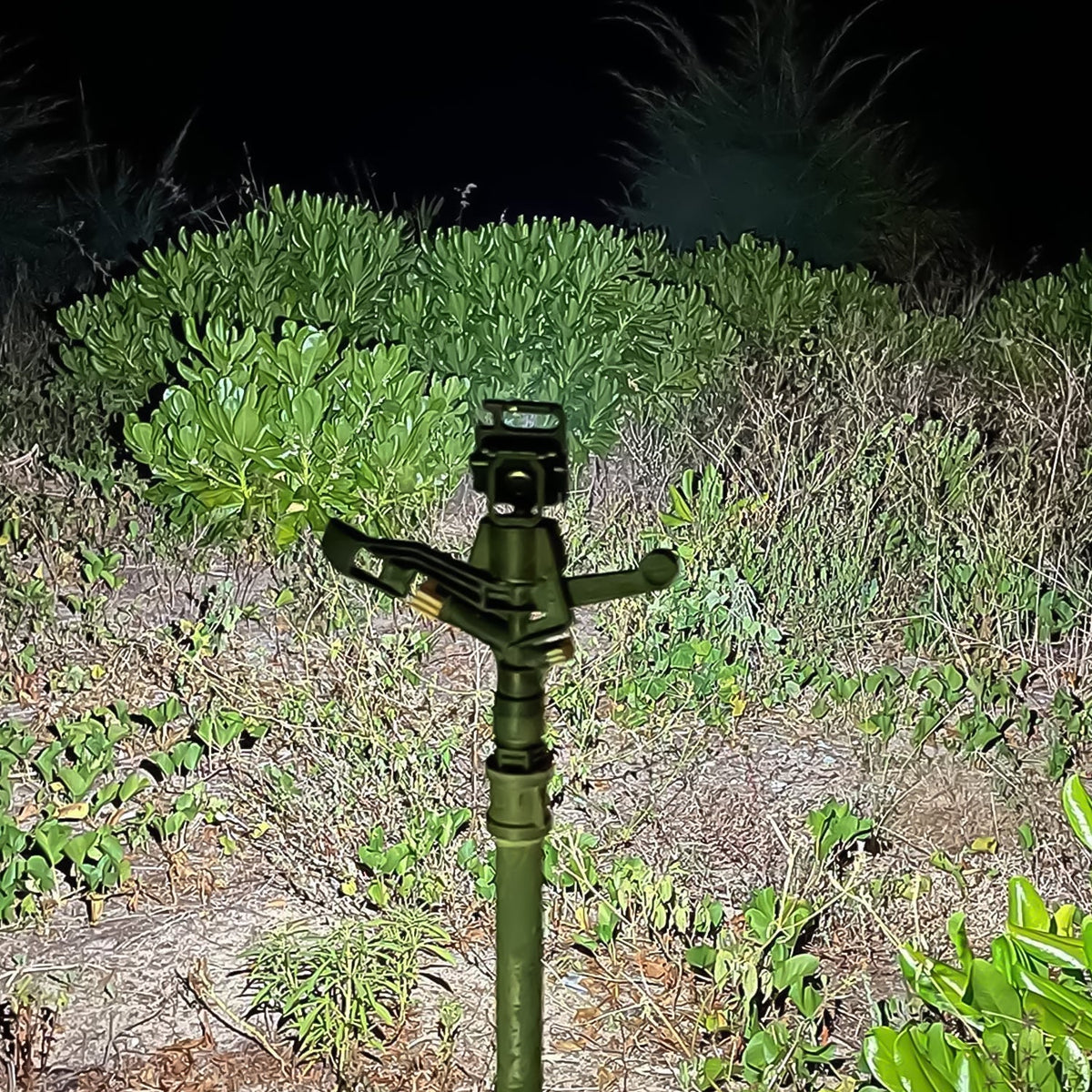 A black mini LED flashlight on a tripod stand, illuminating a dark, leafy outdoor area at night.