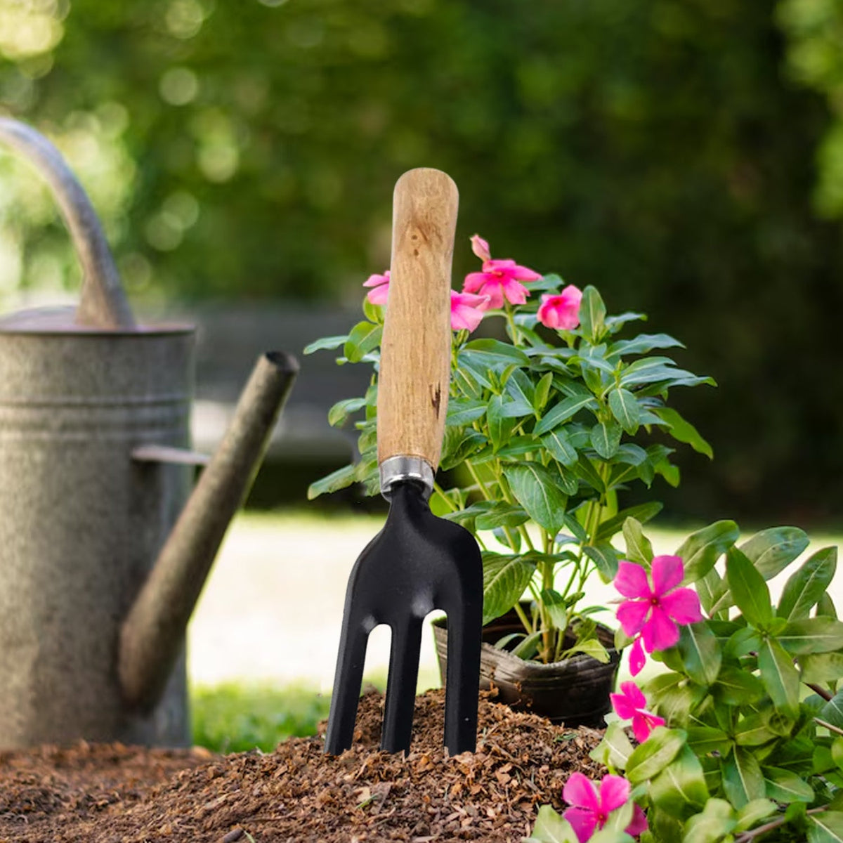 A black three-tine garden fork with a wooden handle stands upright in dark soil next to a pink flowering plant and a