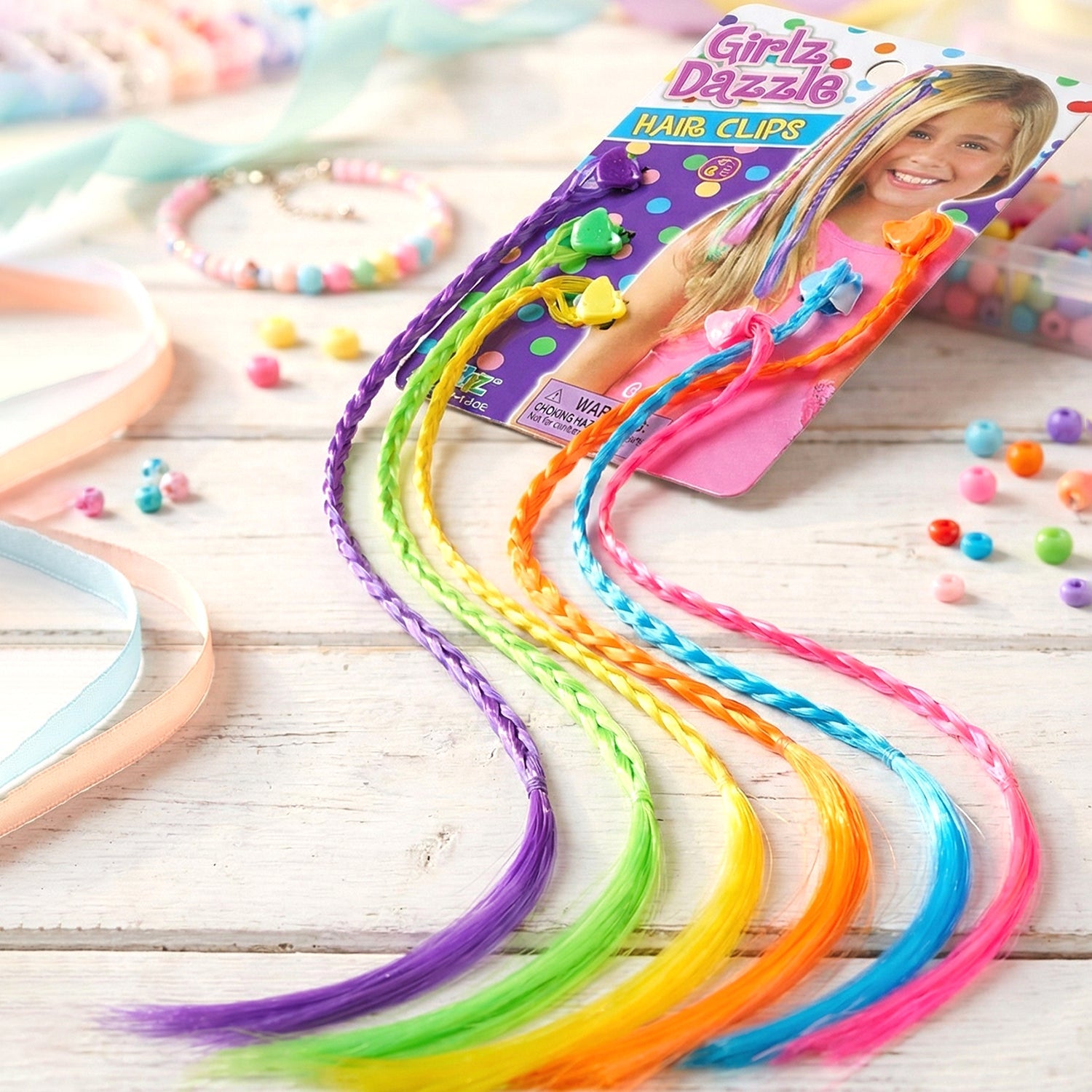 Six colorful braided hair extensions in packaging lie on a rustic white wooden surface with scattered beads and ribbons.