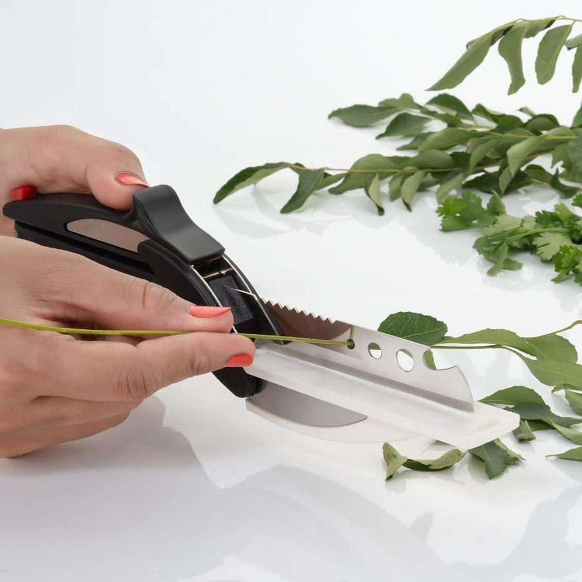 A person's hands use a clever cutter to snip green herbs on a white surface, with more herbs in the background.