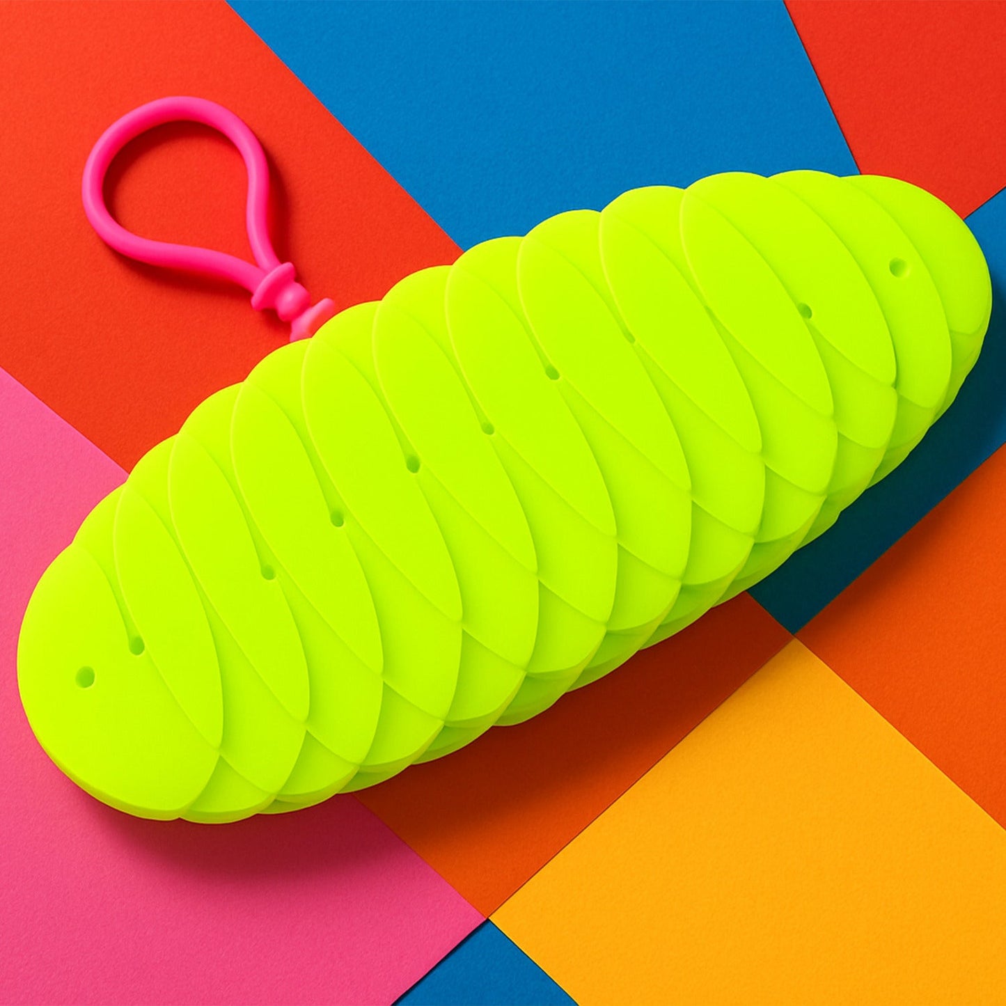 A bright green collapsible silicone ball keychain with a pink loop, shown partially collapsed on a vibrant multi-colored