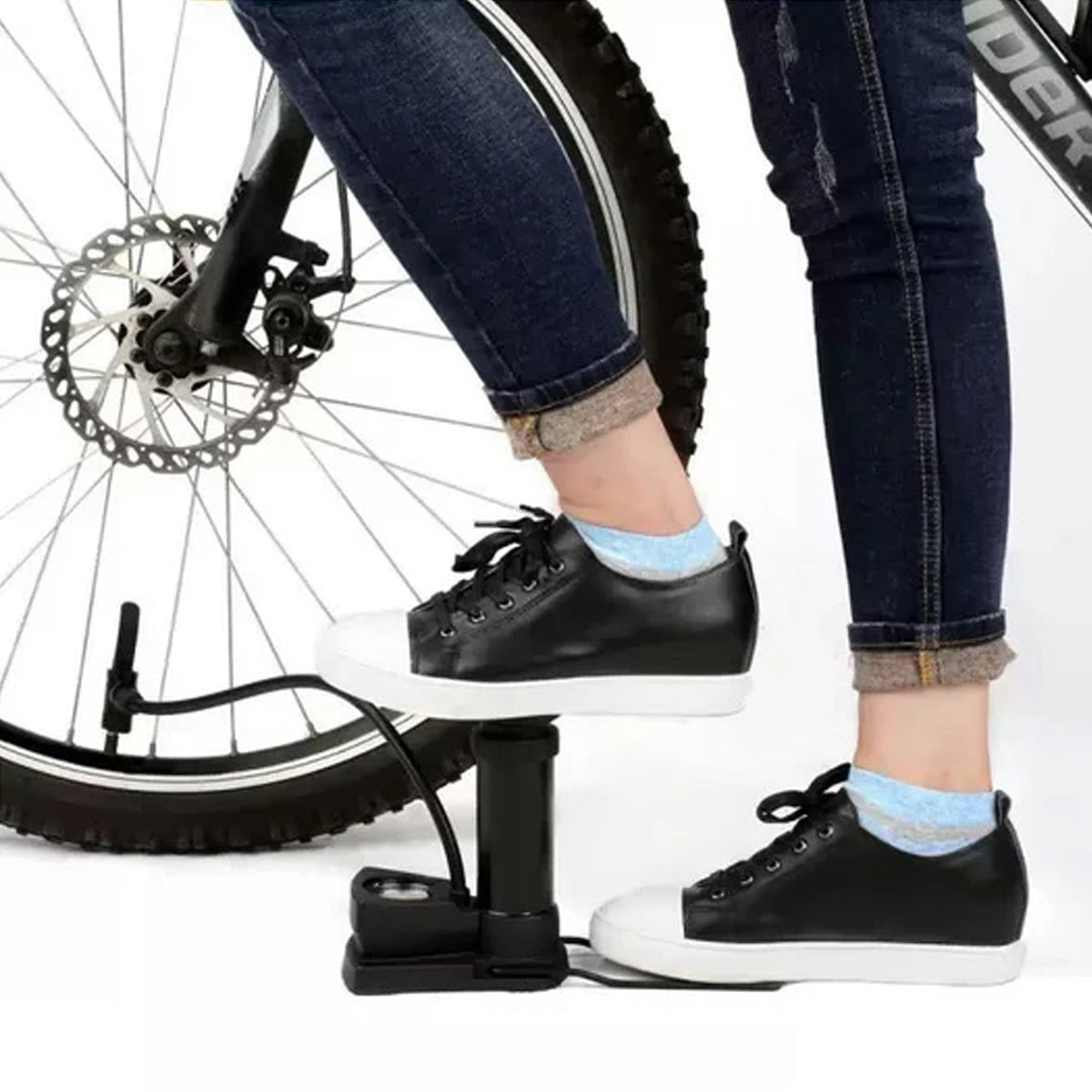 A person in blue jeans and black sneakers uses a black foot pump to inflate a bicycle tire.
