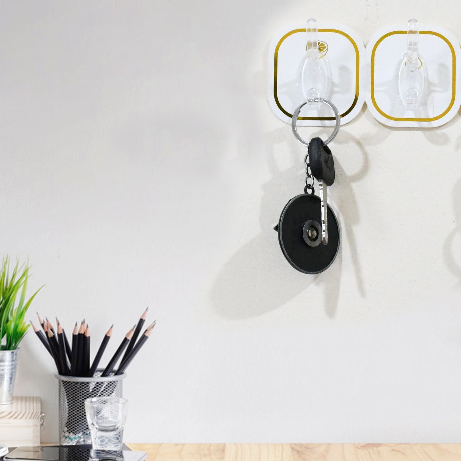 Two transparent adhesive hooks with gold borders on a white wall, holding a set of keys, with a desk and plant below.