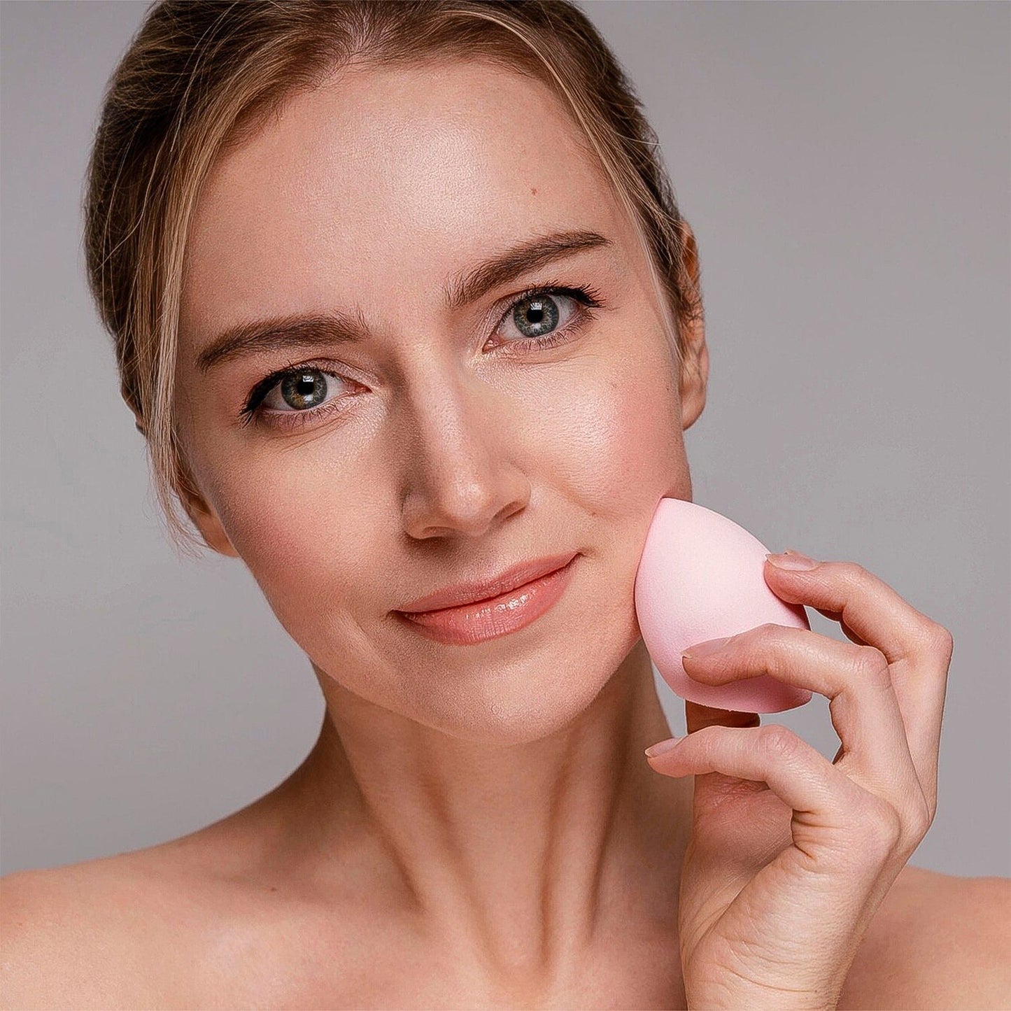 A woman gently dabs her cheek with a light pink, teardrop-shaped makeup sponge, smiling softly.