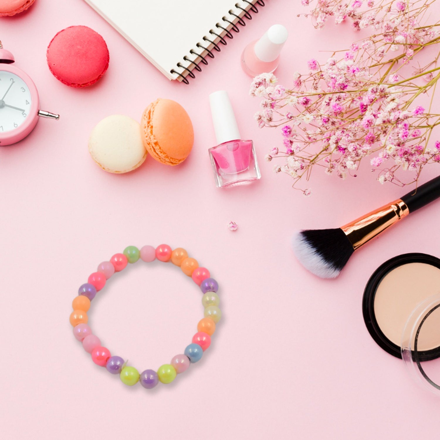 A colorful beaded bracelet is displayed on a pink surface surrounded by macarons, nail polish, a clock, and a makeup brush.