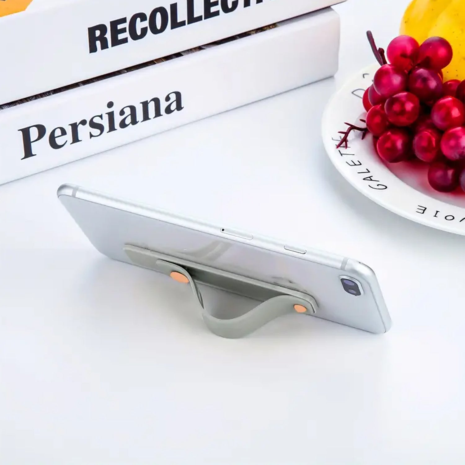 A silver phone propped up horizontally by a gray silicone grip on a white table with books and grapes.
