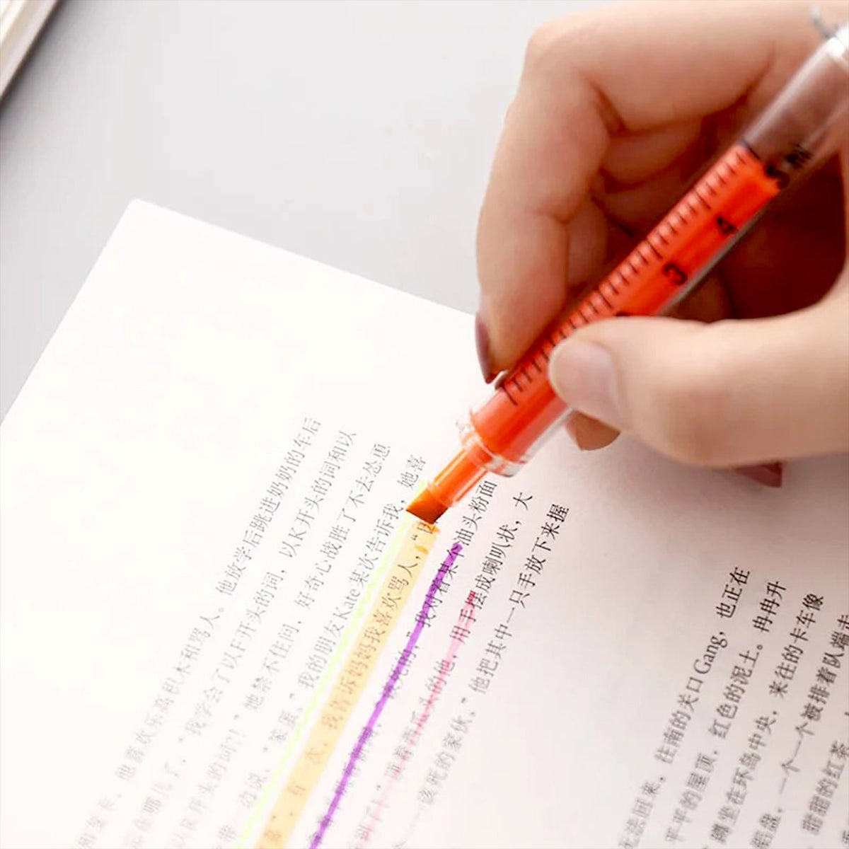 A hand highlights text in a book with an orange syringe highlighter, showing the pen in action.