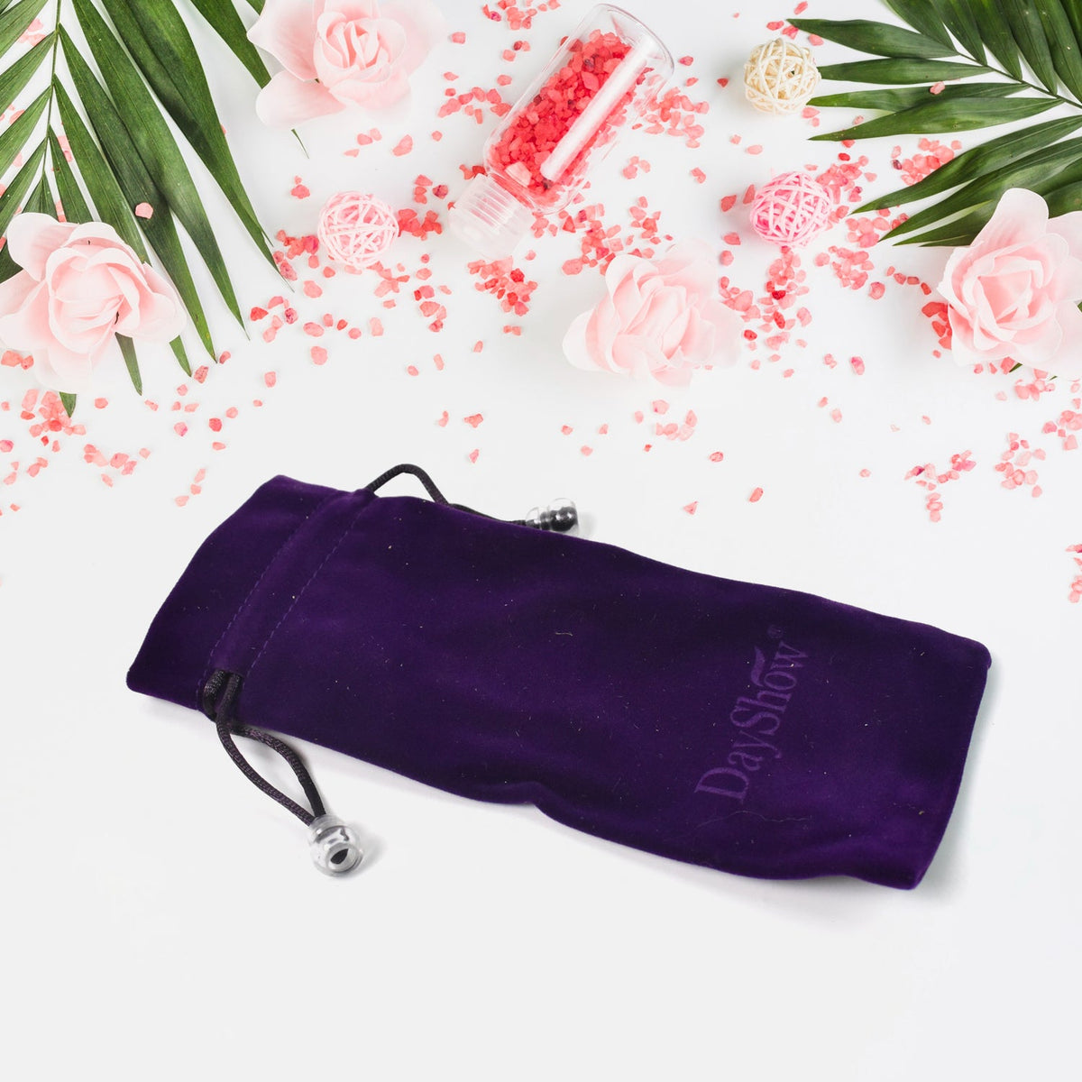 A purple velvet drawstring bag with a DayShow logo rests on a white surface with pink flowers and scattered red beads.