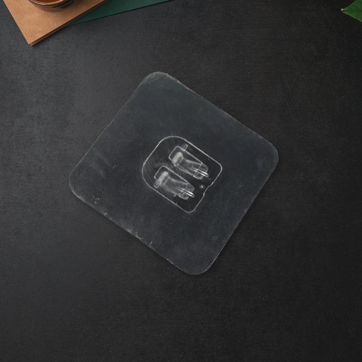 A clear self-adhesive wall hook with two prongs is centered on a dark grey surface, viewed from above.
