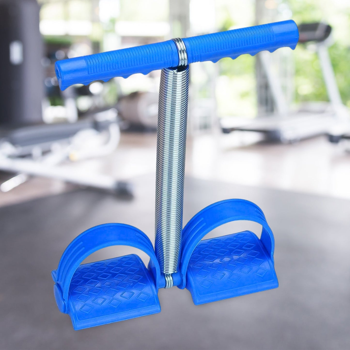 A blue foot pedal exerciser with a metal spring stands upright on a dark floor in a blurred gym setting.