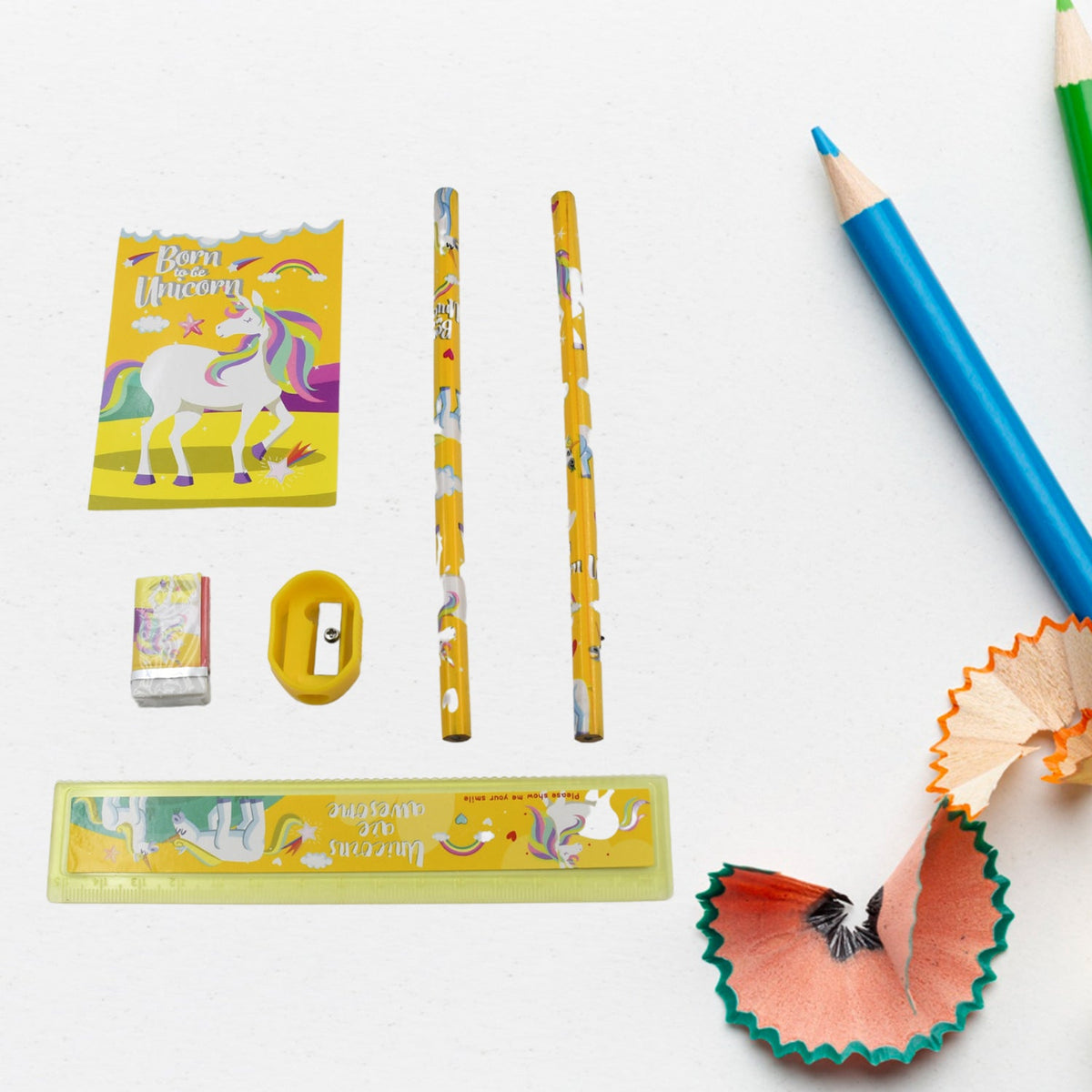 Unicorn-themed pencils, ruler, sharpener, eraser, and notebook arranged on a white surface with colored pencil shavings.