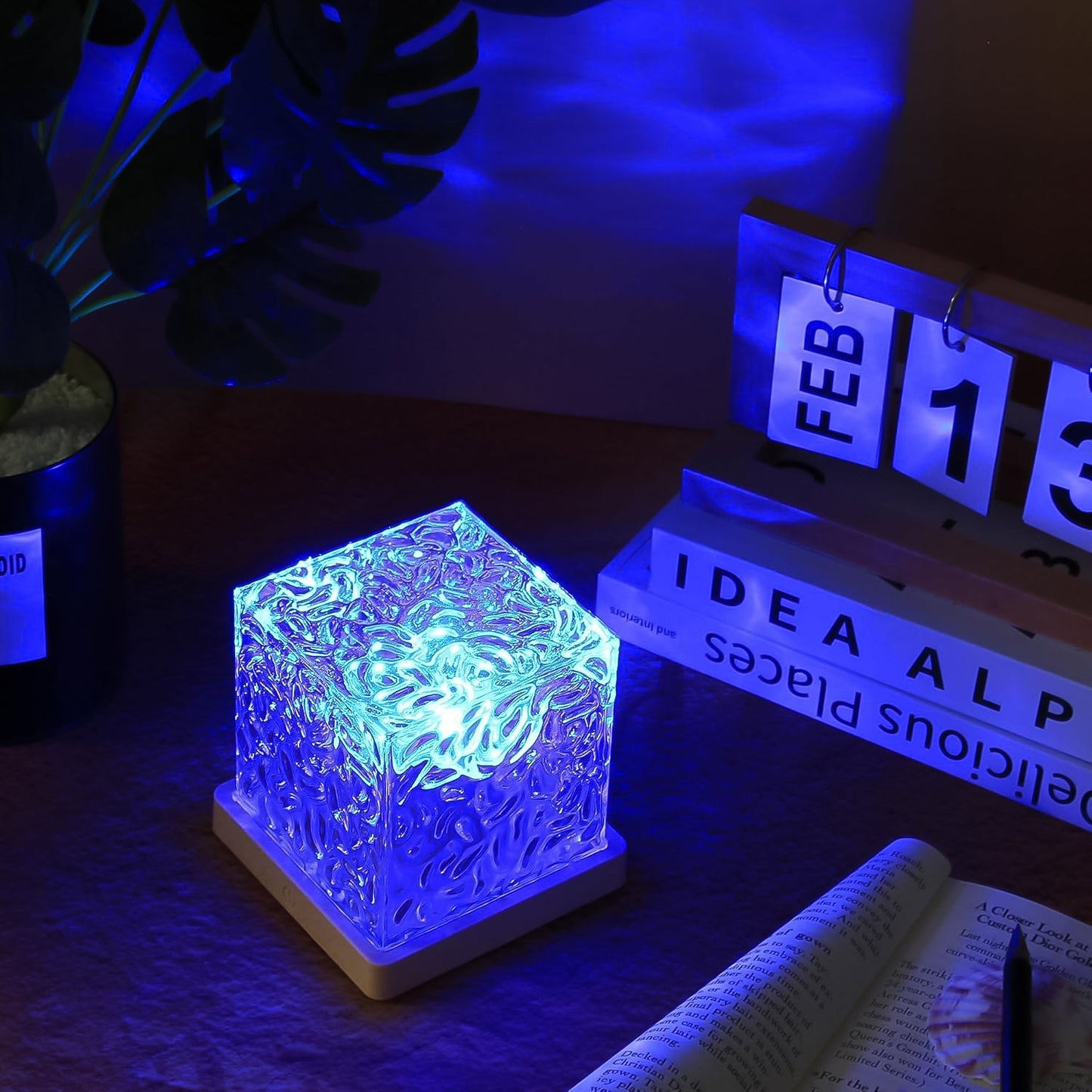 A crystal cube lamp projecting blue ripples on a desk with a calendar, books, and a plant in the background.