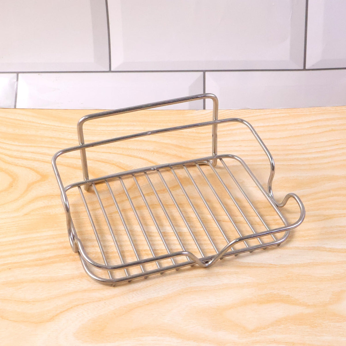A stainless steel wire soap dish rests on a light wood surface with white subway tiles in the background.