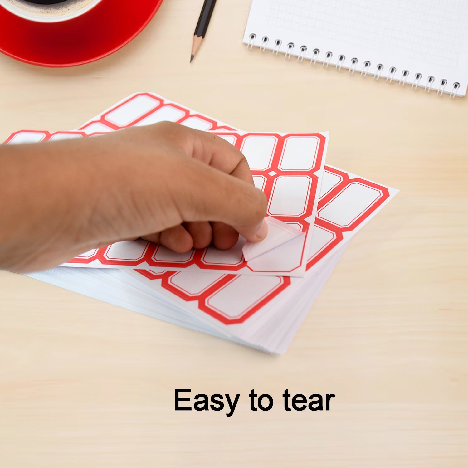 A hand peeling a red-bordered label from a sheet on a wooden desk, with a red cup and notebook.