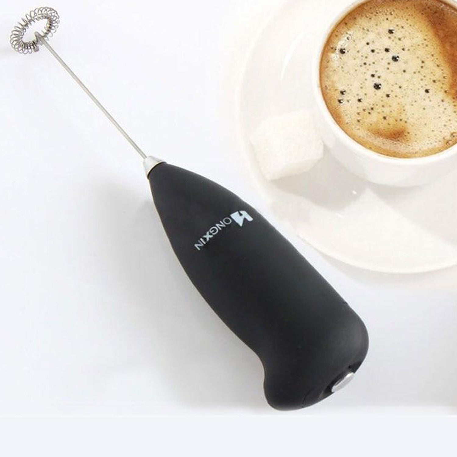 A black frother with a silver whisk rests on a white surface next to a cup of coffee and sugar cube.