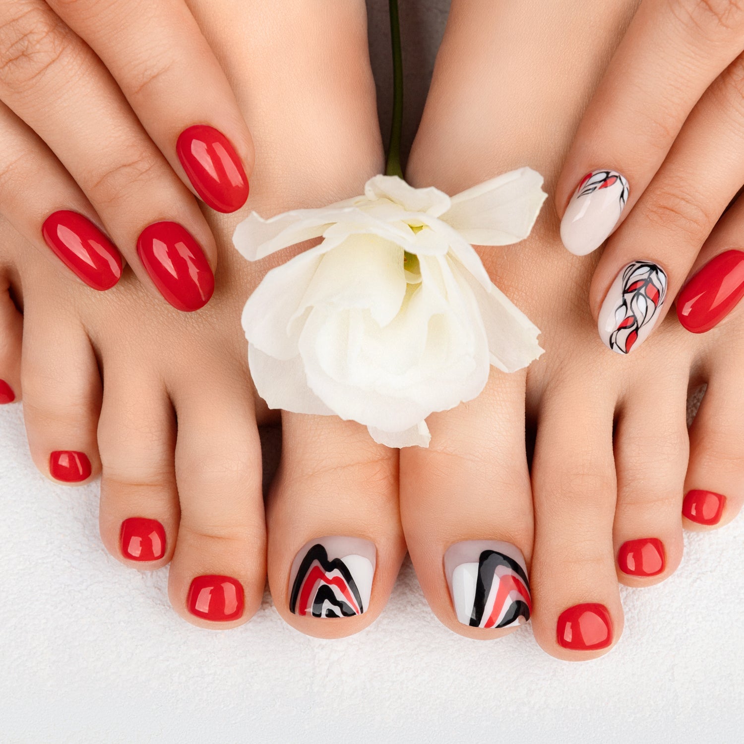 Manicured hands and feet with red and patterned nail polish holding a white flower.