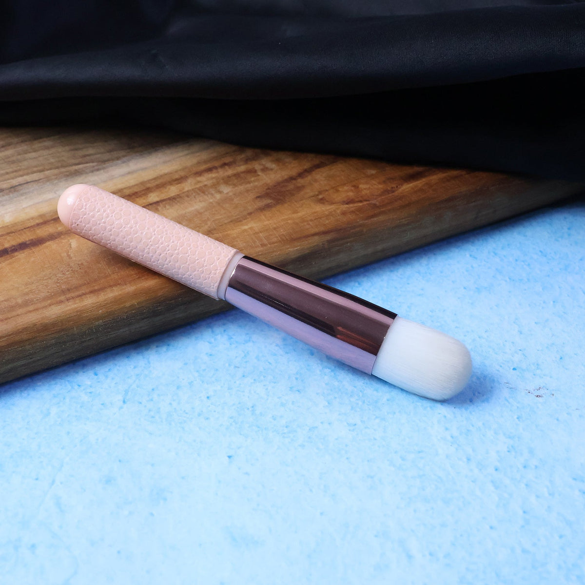 A pink and rose gold makeup brush on a blue textured surface with a wooden board and black fabric.
