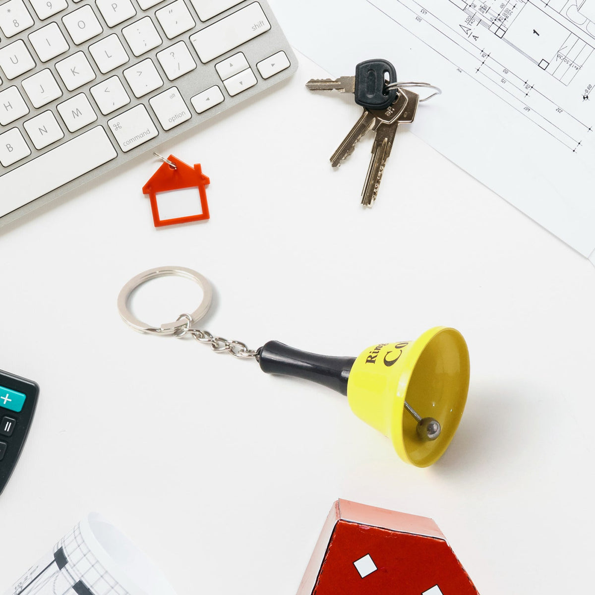 A yellow mini hand bell keychain with the phrase "Ring for a Coffee" on a white desk with keys, a keyboard, and a calculator.