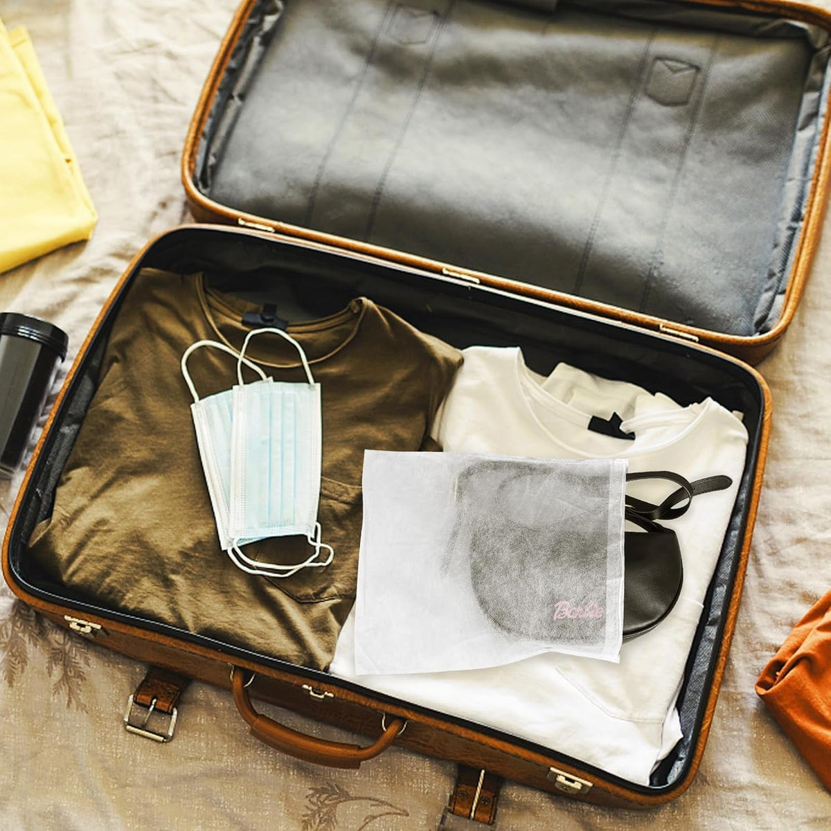 An open suitcase with folded clothes, two face masks, and a white non-woven bag containing a black item.